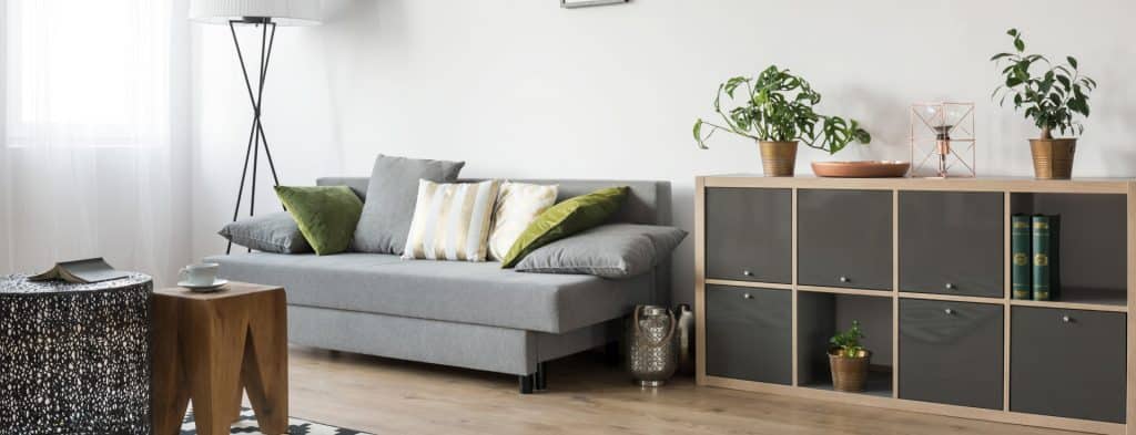 Minimalist living room featuring a gray fabric sofa with green and striped cushions, a wooden cubby storage unit, potted plants, and a decorative lamp on light wood flooring